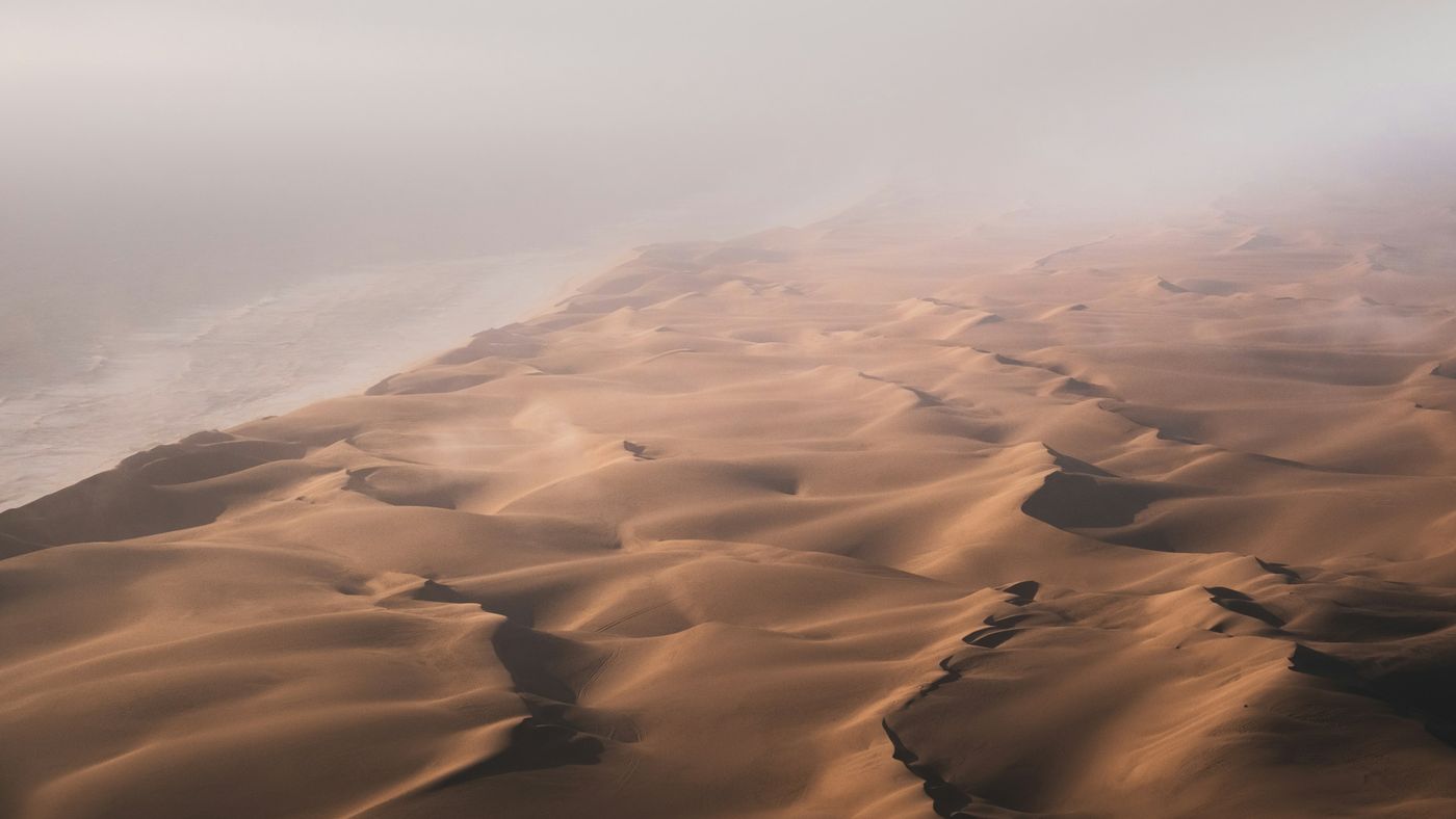 Vue aérienne d’un champ de dunes qui se perd dans la brume, ombres longues sur le sable.