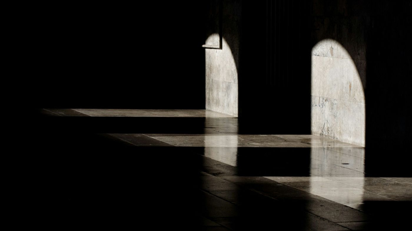 Deux arches de lumière rasante sur un mur de pierre, dans un intérieur plongé dans l’ombre.