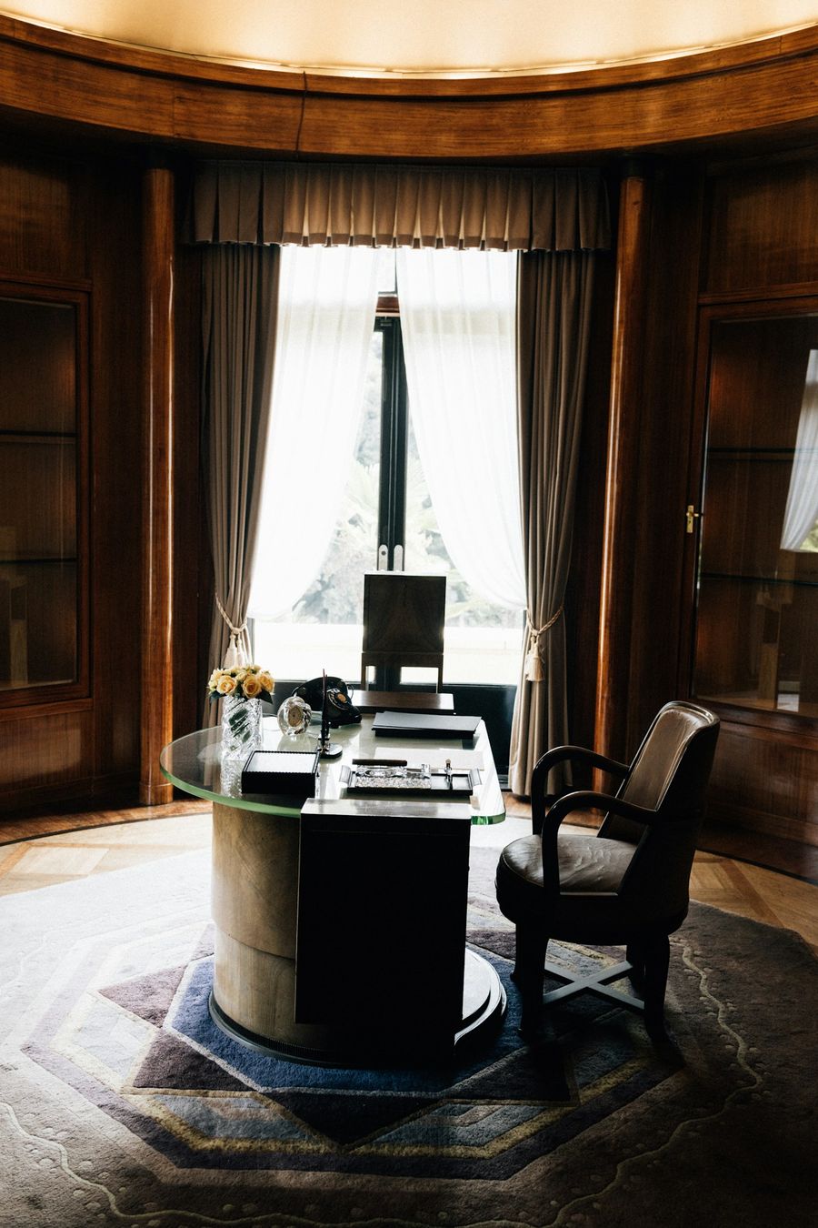 Bureau aux boiseries sombres, dossiers fermés sur la table, haute fenêtre voilée de rideaux, pièce vide.