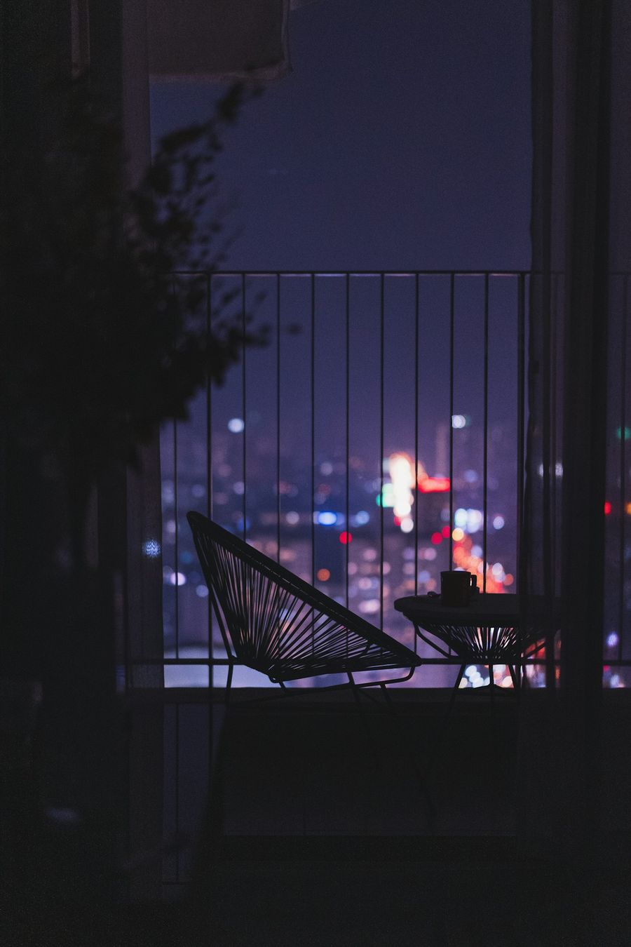 La nuit, un fauteuil et une petite table en silhouette devant les lumières floues d’une ville lointaine.